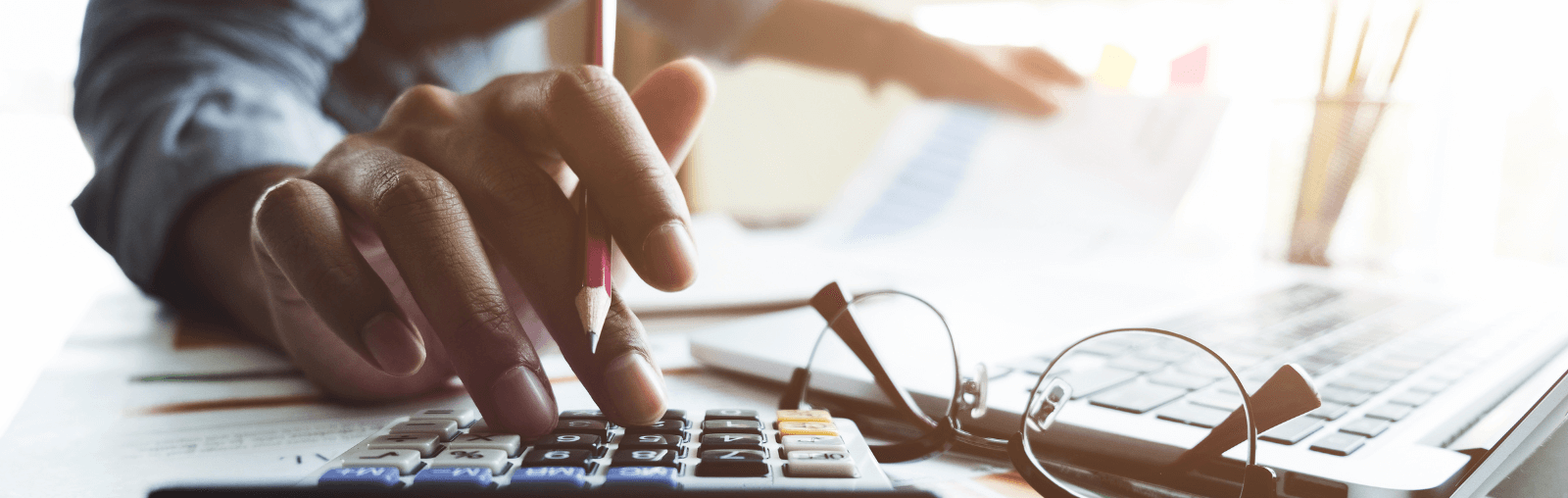 Close-up of a hand using a calculator, with a pencil, eyeglasses, and a laptop keyboard visible.